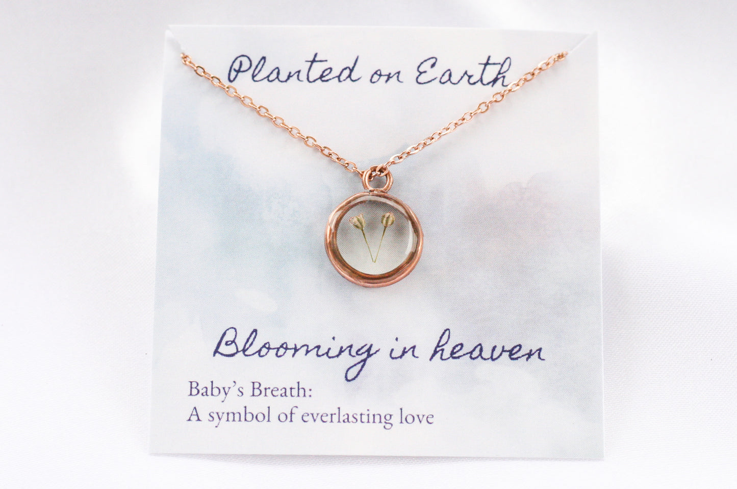 two pieces of babys breath in a rose gold pendant necklace on a card reading "planted on earth blooming in heaven"