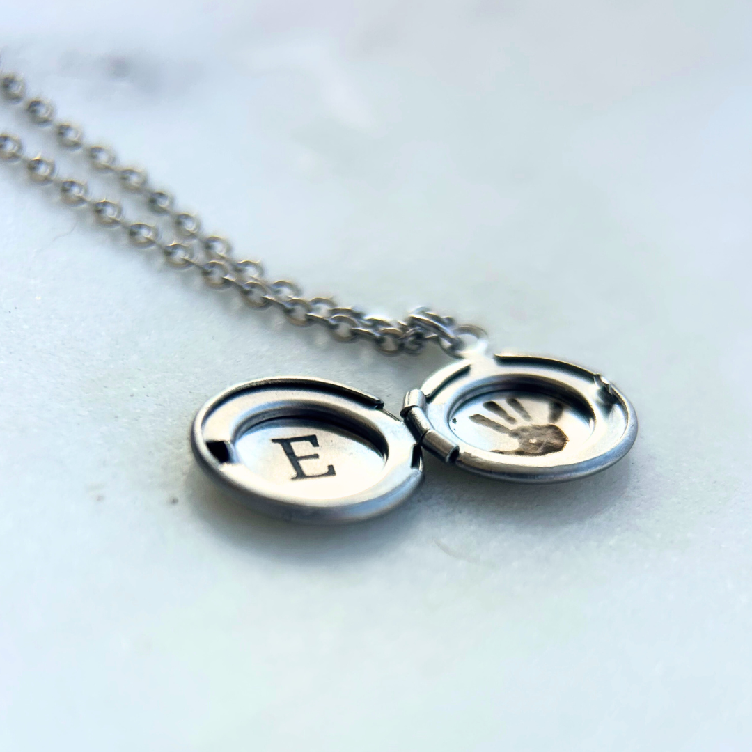 Rest of the Nest silver necklace with a locket featuring the letter 'E' and a personalized engraved handprint on a light background 