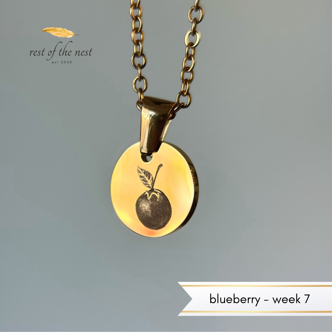 Rest of the Nest Gold necklace with a blueberry pendant on a gray background.