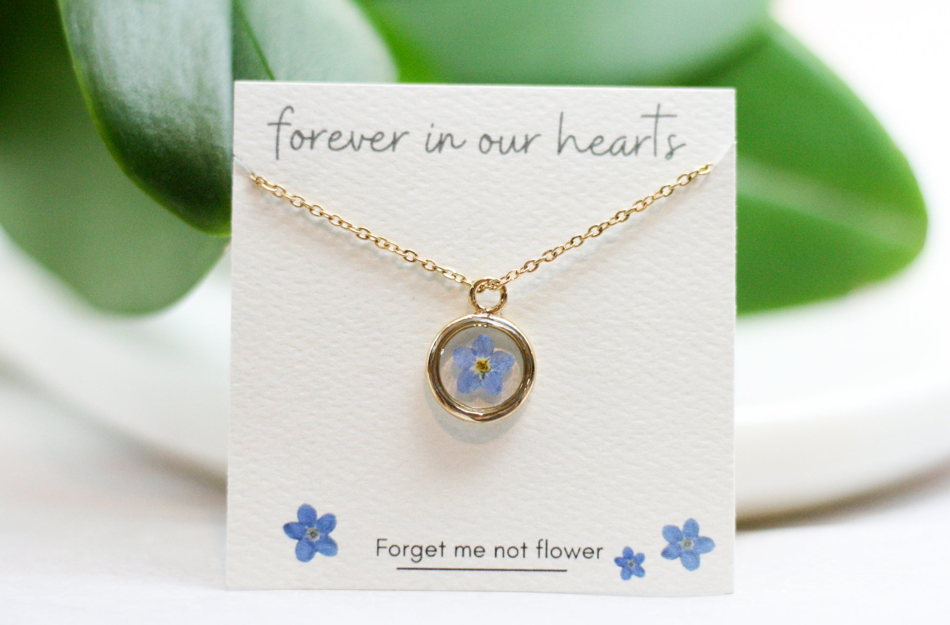 gold forget me not necklace in a gold pendant on a card reading "forever in our hearts" at the top and "forget me not flower" at the bottom next to hand drawn forget me not flowers