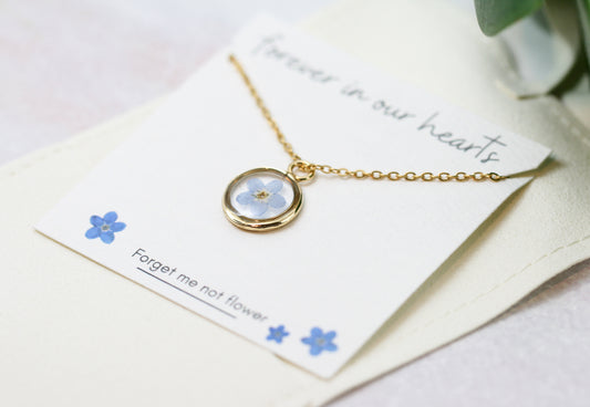 gold forget me not necklace in a gold pendant on a card reading "forever in our hearts" at the top and "forget me not flower" at the bottom next to hand drawn forget me not flowers