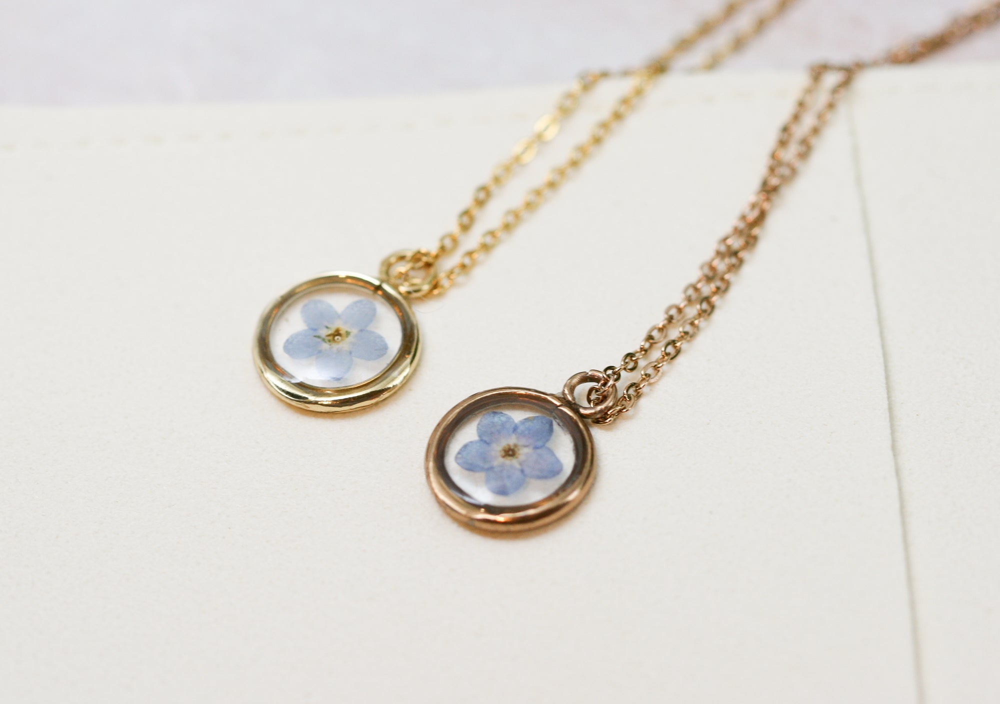 gold and rose gold necklaces with a blue forget me not flower inside resting on an off white microfiber necklace bag