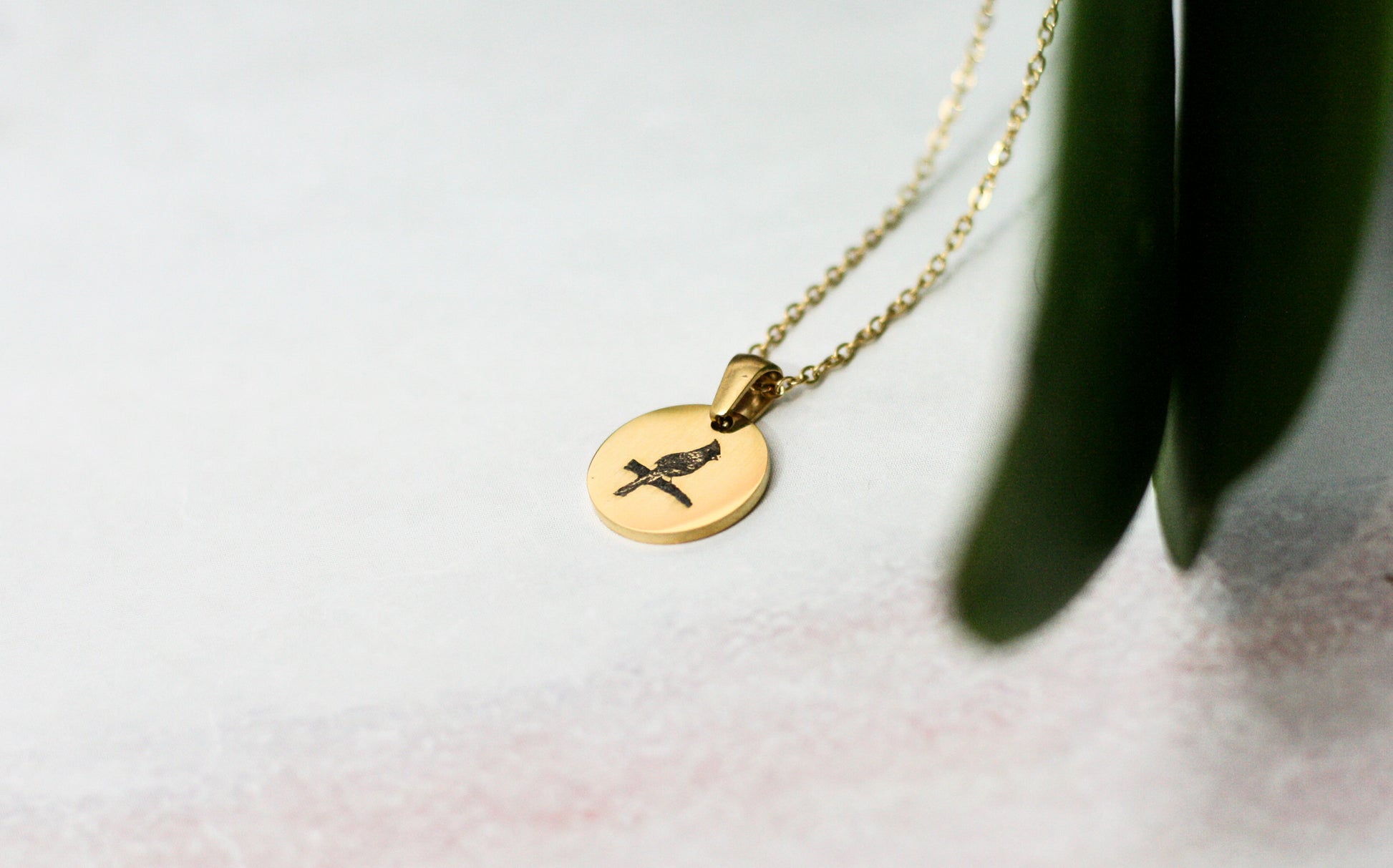 gold cardinal pendant necklace next to a green leaf