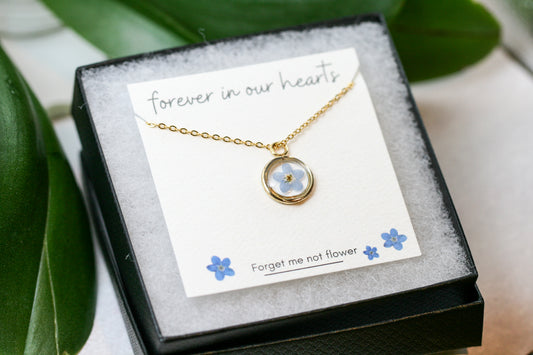 gold forget me not necklace in a gold pendant on a card reading "forever in our hearts" at the top and "forget me not flower" at the bottom next to hand drawn forget me not flowers in a black necklace box