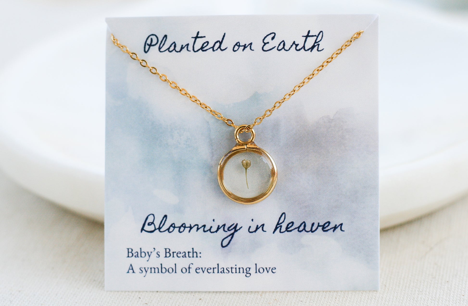 single piece of babys breath inside a gold pendant necklace on a card reading "planted on earth blooming in heaven"