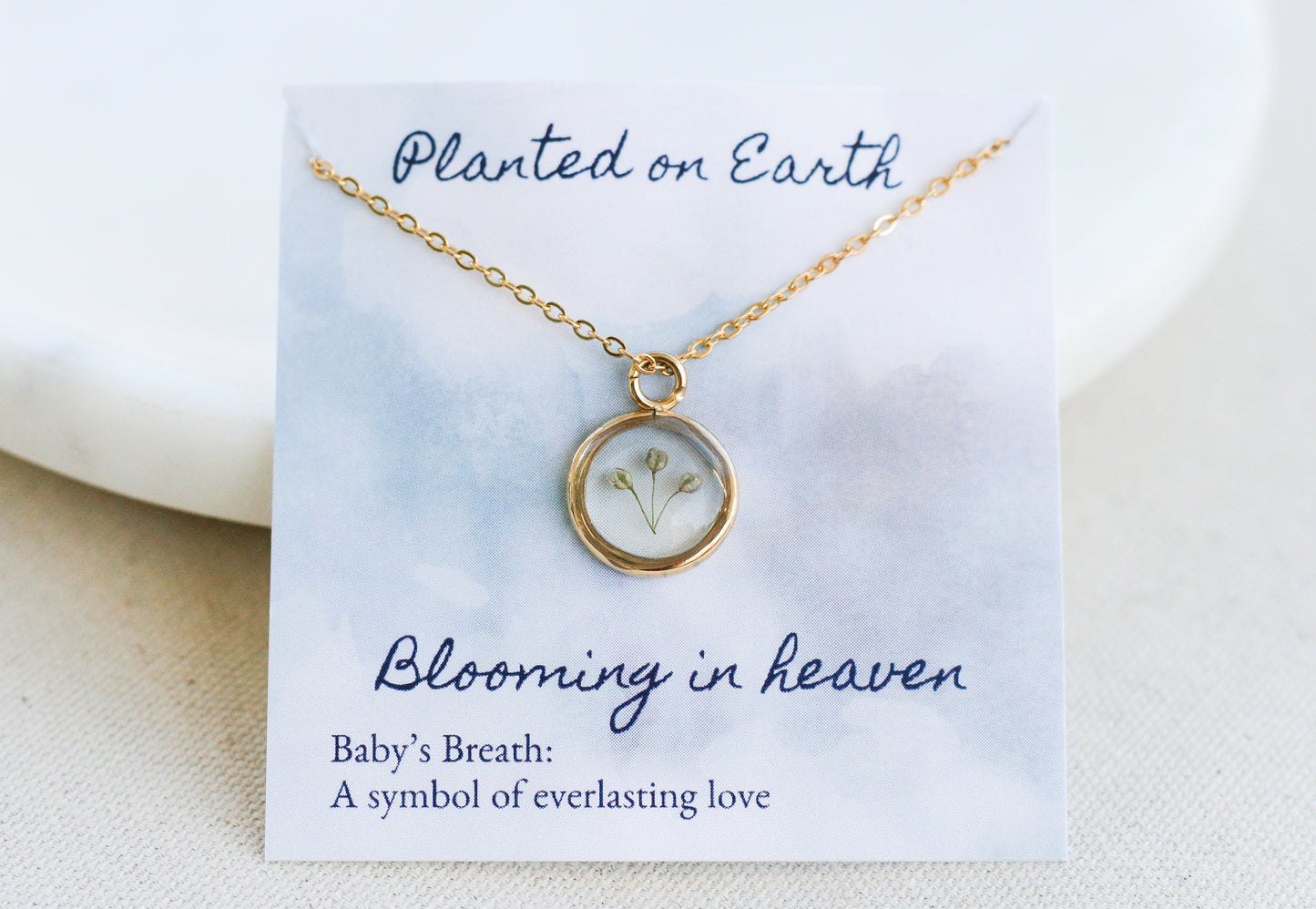 three pieces of babys breath inside a gold necklace pendant on top of a card reading "planted on earth blooming in heaven" and "babys breath: a symbol of everlasting love" at the bottom