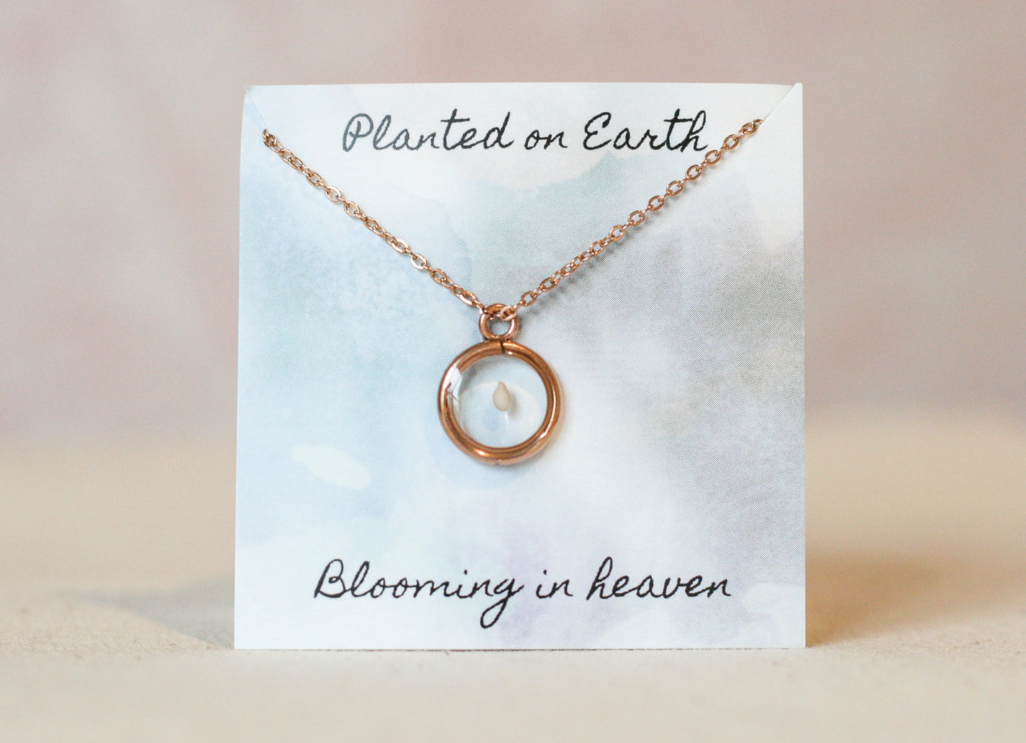 one sesame seed inside of a rose gold necklace pendant on a card reading "planted on earth blooming in heaven"